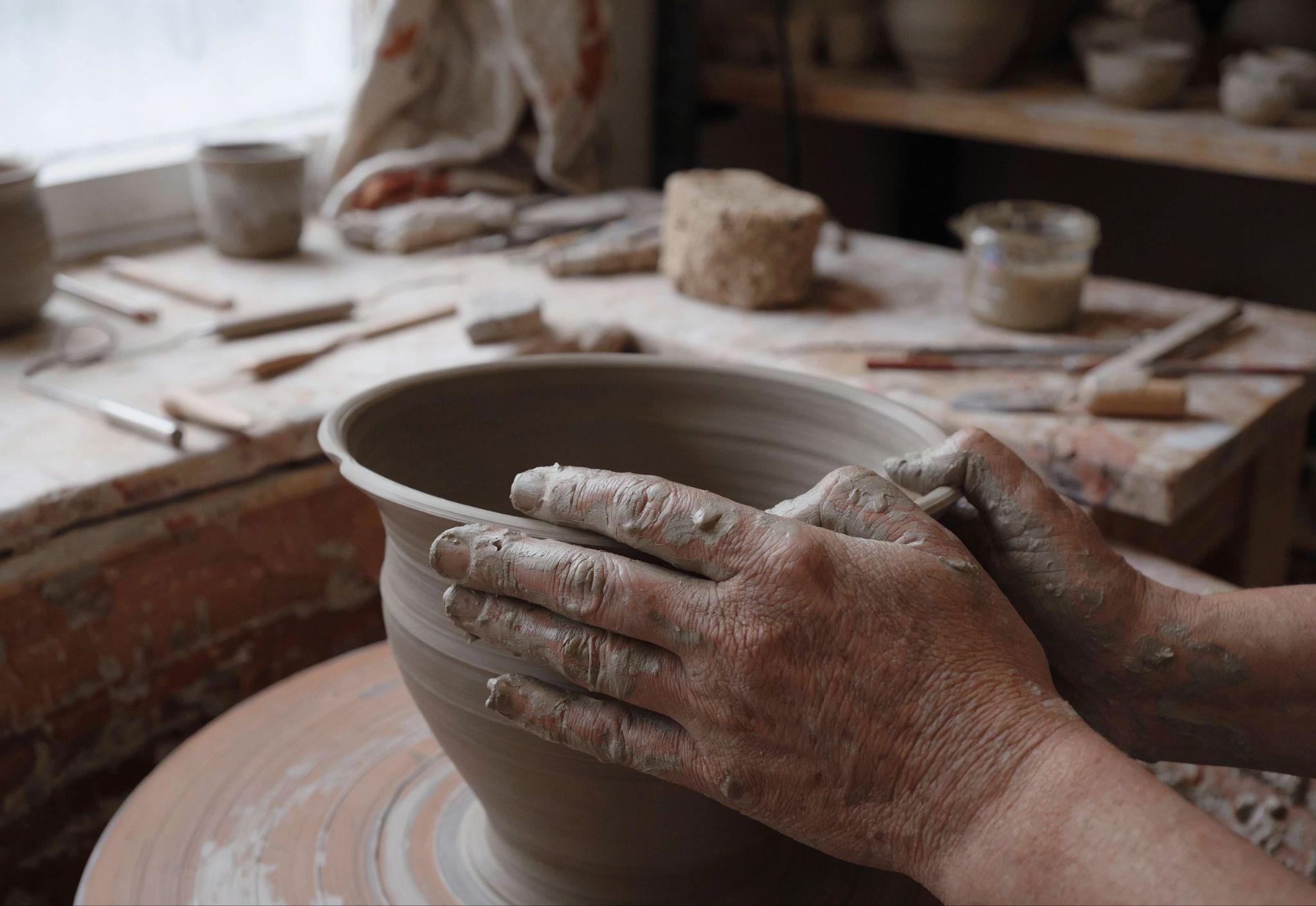Close-up of a ceramic artist's clay-covered hands shaping a bowl in a cluttered home studio