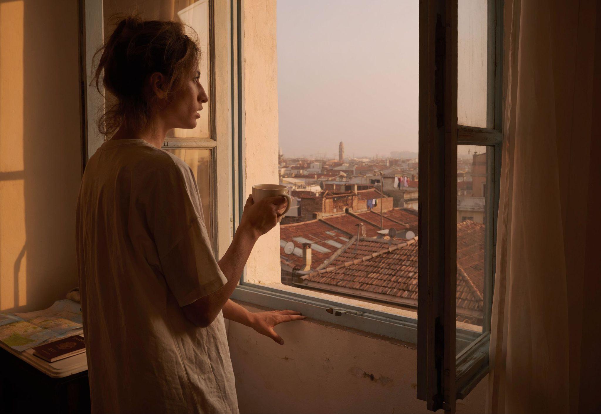 Woman holding coffee by an open window overlooking unfamiliar rooftops in the morning light